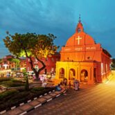 Christ Church at Dutch Square in Malacca, Malaysia, illuminated at night with colorful trishaws and visitors in the foreground.