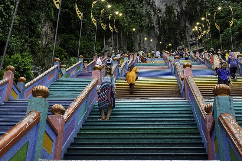 Tourist wearing a sarong posing for a photo on the vibrant Batu Caves rainbow stairs, surrounded by other visitors and decorative lighting poles.