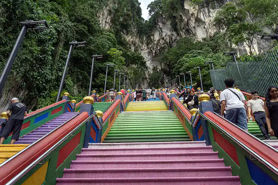 Visitors climbing the colourful 272 rainbow steps at Batu Caves, with the limestone cliff and cave entrance visible at the top