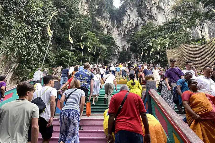  Crowds of tourists and devotees climbing the rainbow staircase at Batu Caves during peak midday hours, with the limestone cliff and Temple Cave entrance visible ahead