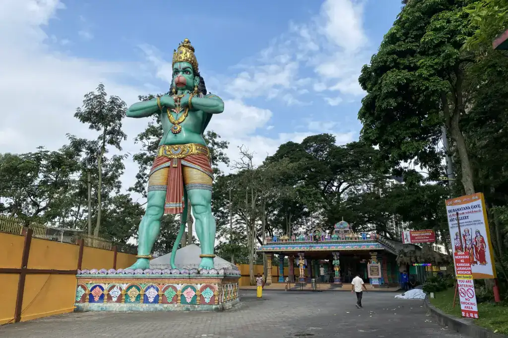 The large green Hanuman statue standing at the entrance to the Ramayana Cave at Batu Caves, with the ticket counter and colourful gopuram visible behind it