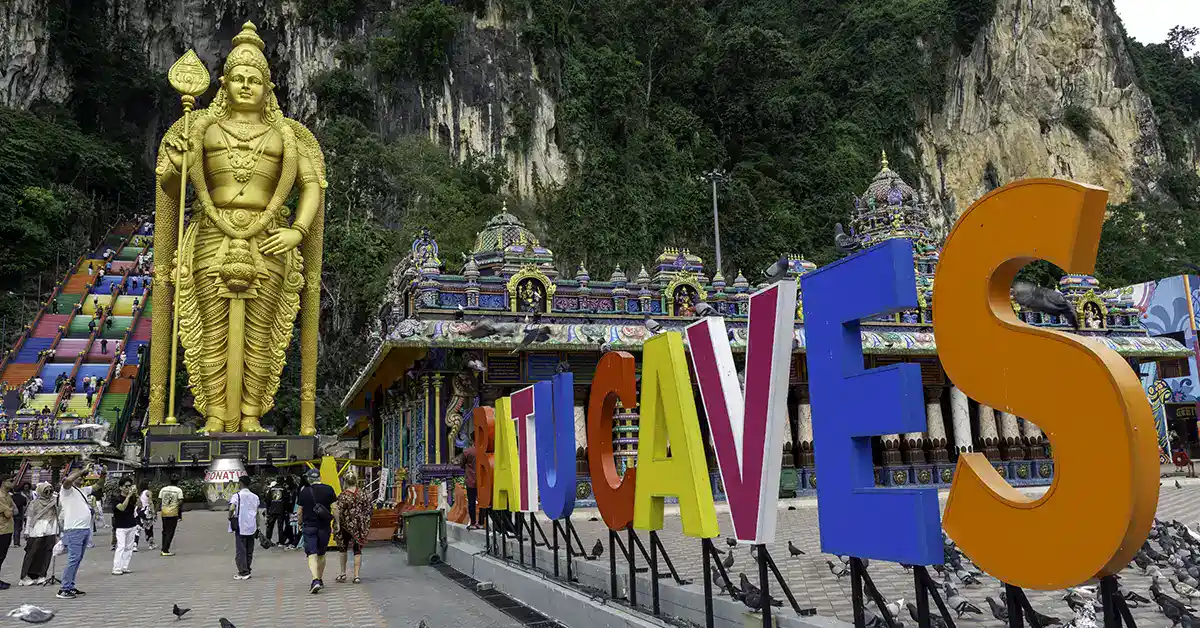 Tourists exploring Batu Caves courtyard with the golden Lord Murugan statue, rainbow staircase, and colourful sign in Kuala Lumpur