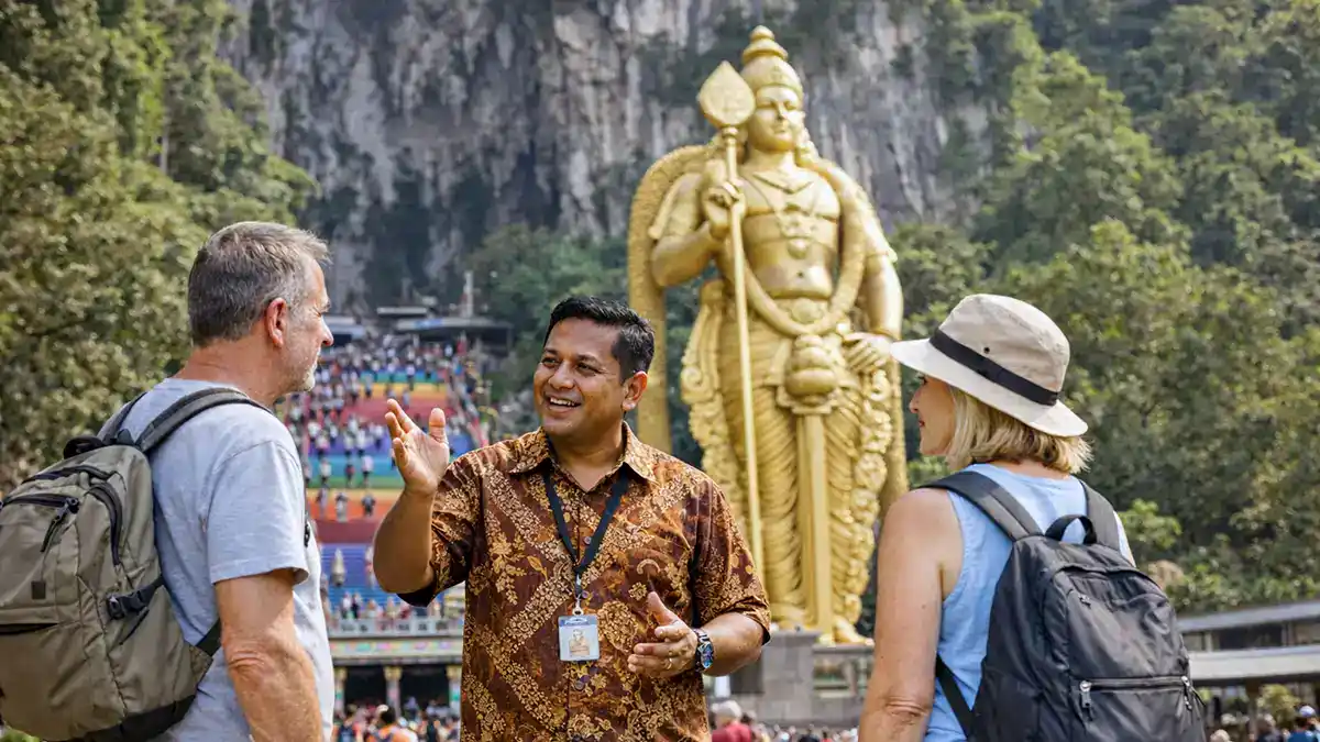 A local tour guide explaining the history of Batu Caves to two tourists, with the golden Lord Murugan statue and rainbow staircase in the background