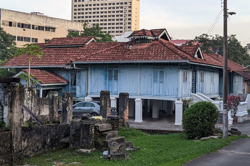 Traditional blue Malay wooden house with red tile roof in Kampung Baru with a modern hotel tower rising directly behind it — things to do in Kampung Baru KL include walking through this living Malay village inside the city