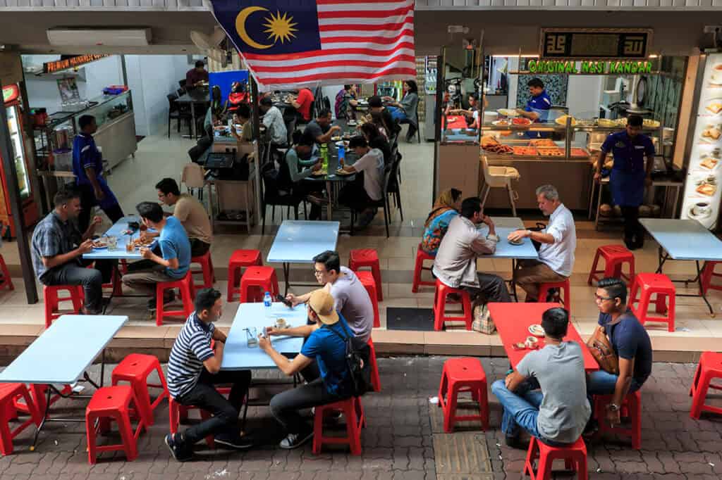Locals from different communities eating and socialising at open-air plastic tables outside a mamak stall in Kuala Lumpur with a Malaysian flag flying above