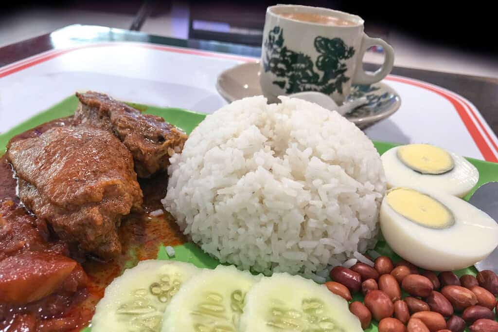 Nasi Lemak served on a green plate at a Kuala Lumpur kopitiam — coconut rice with rendang, sambal, boiled egg, peanuts and cucumber with a traditional kopi cup in the background
