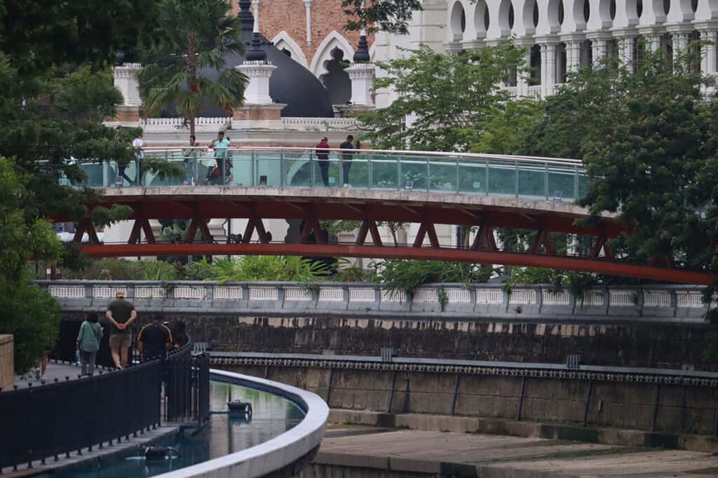Tourists walking along the River of Life elevated walkway beside the Klang River, with the Sultan Abdul Samad Building visible in the background — one of the best free things to do in Kuala Lumpur in the Merdeka Square zone