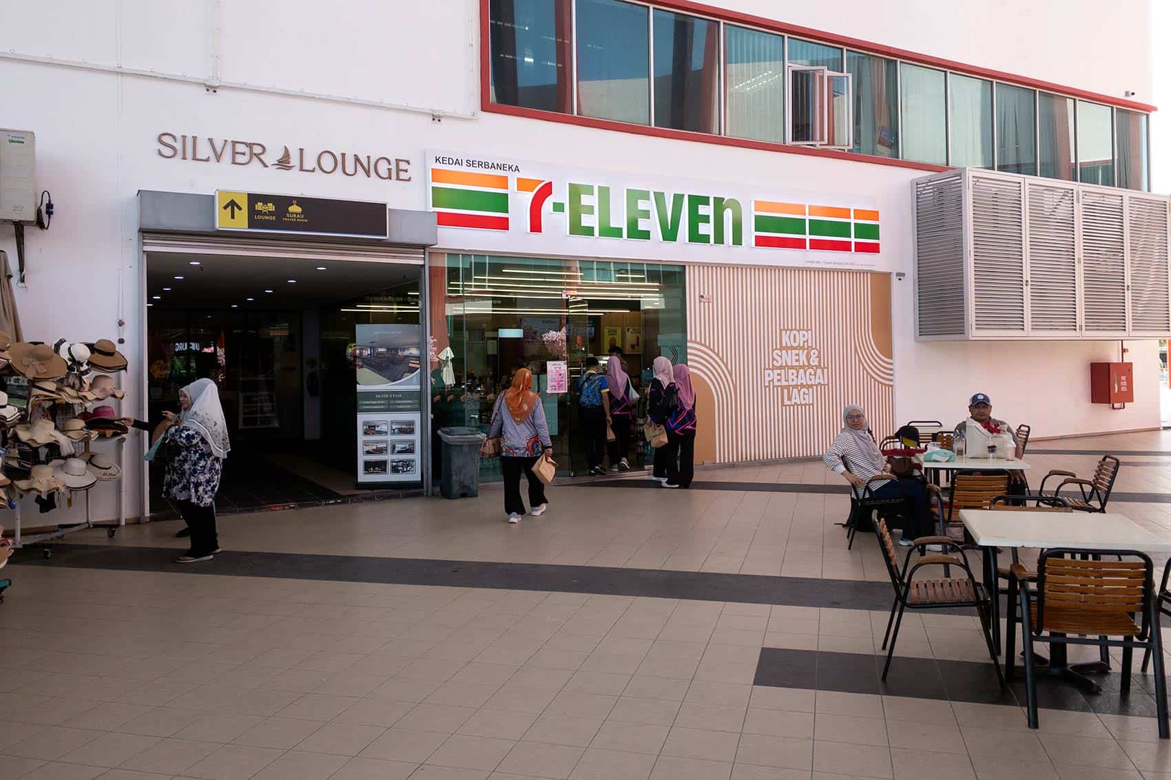 Marina Island Jetty terminal showing Silver Lounge entrance with upward arrow sign, 7-Eleven convenience store, outdoor seating, and hat vendors — facilities available before boarding the Pangkor Island ferry