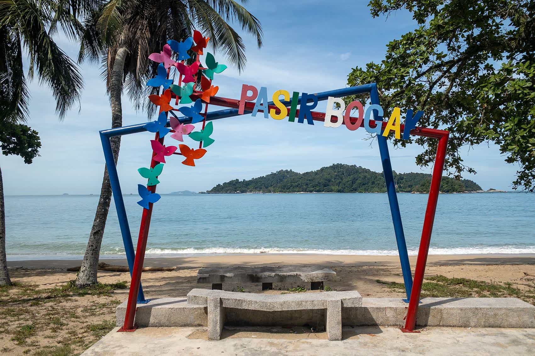 Colourful Pasir Bogak arch sign on the beach at Pangkor Island, with Pangkor Laut island visible in the background.