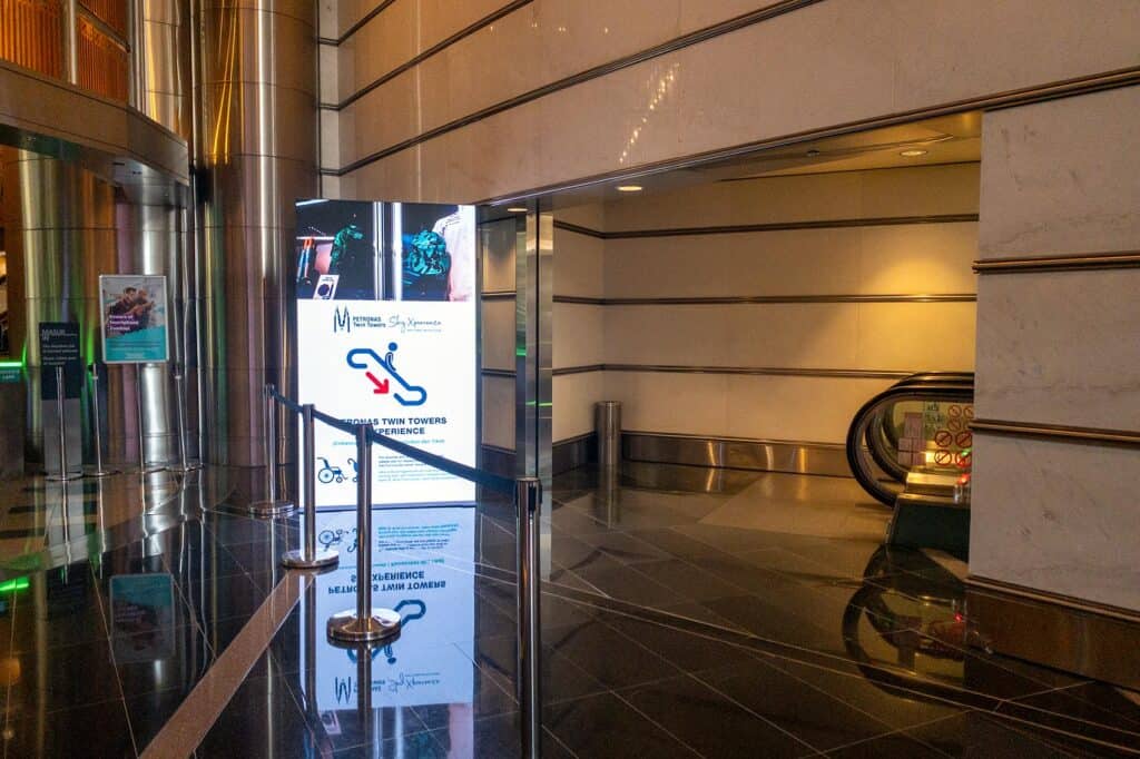 Petronas Twin Towers Sky Xperience entrance signage with escalator leading down to Daftar Masuk check-in area
