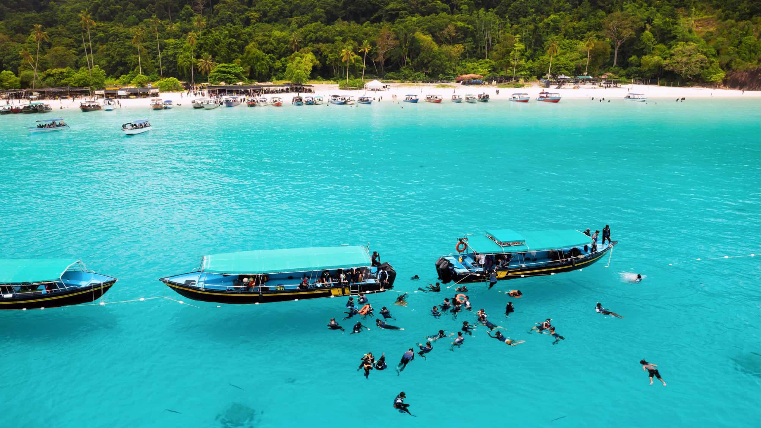 Aerial view of snorkellers and wooden boats in turquoise water off a white sand beach with rainforest — places to visit in Malaysia