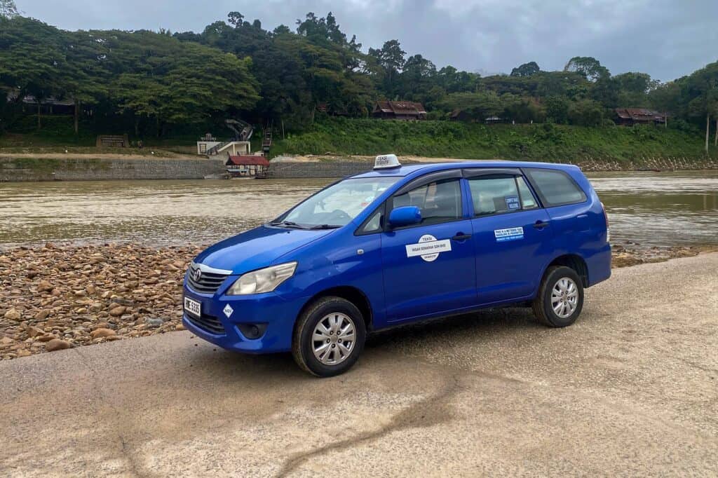 Blue Toyota Innova taxi parked at the Kuala Tahan riverside landing — private transfer arrival point for Taman Negara visitors from Kuala Lumpur