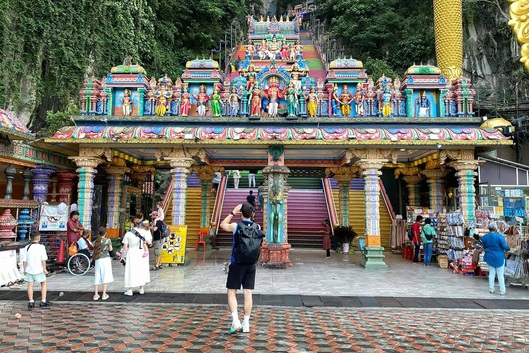 The entrance gate and forecourt of Sri Subramaniar Swamy Temple at Batu Caves — the dress code information board (yellow sign, centre) is visible at the point where temple staff check visitor clothing before allowing them to climb the 272-step rainbow staircase to the Temple Cave