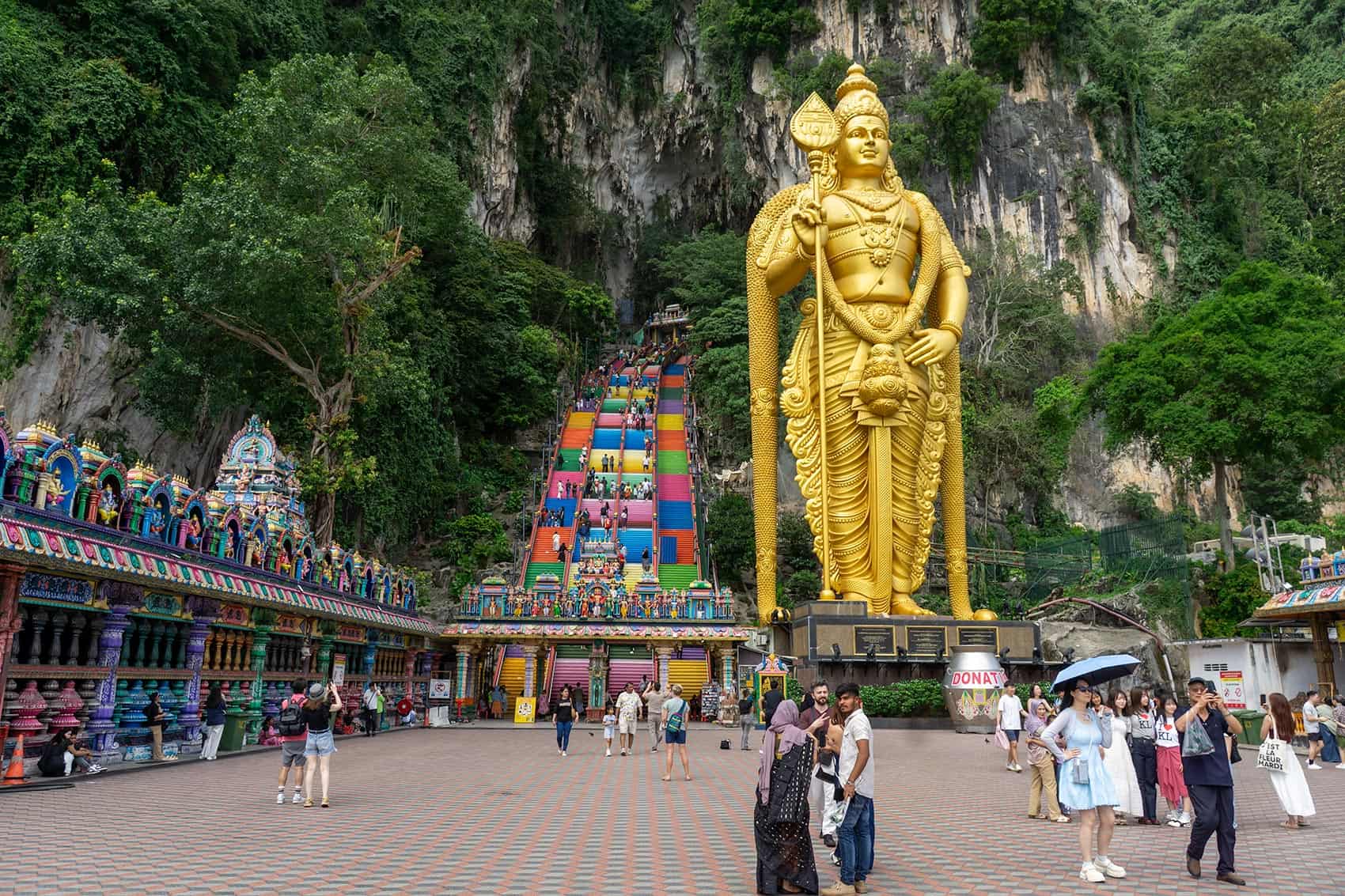 The Batu Caves forecourt with the 42.7-metre golden Lord Murugan statue and the rainbow staircase in the background — visitors in casual clothing including shorts visible at ground level where no dress code applies