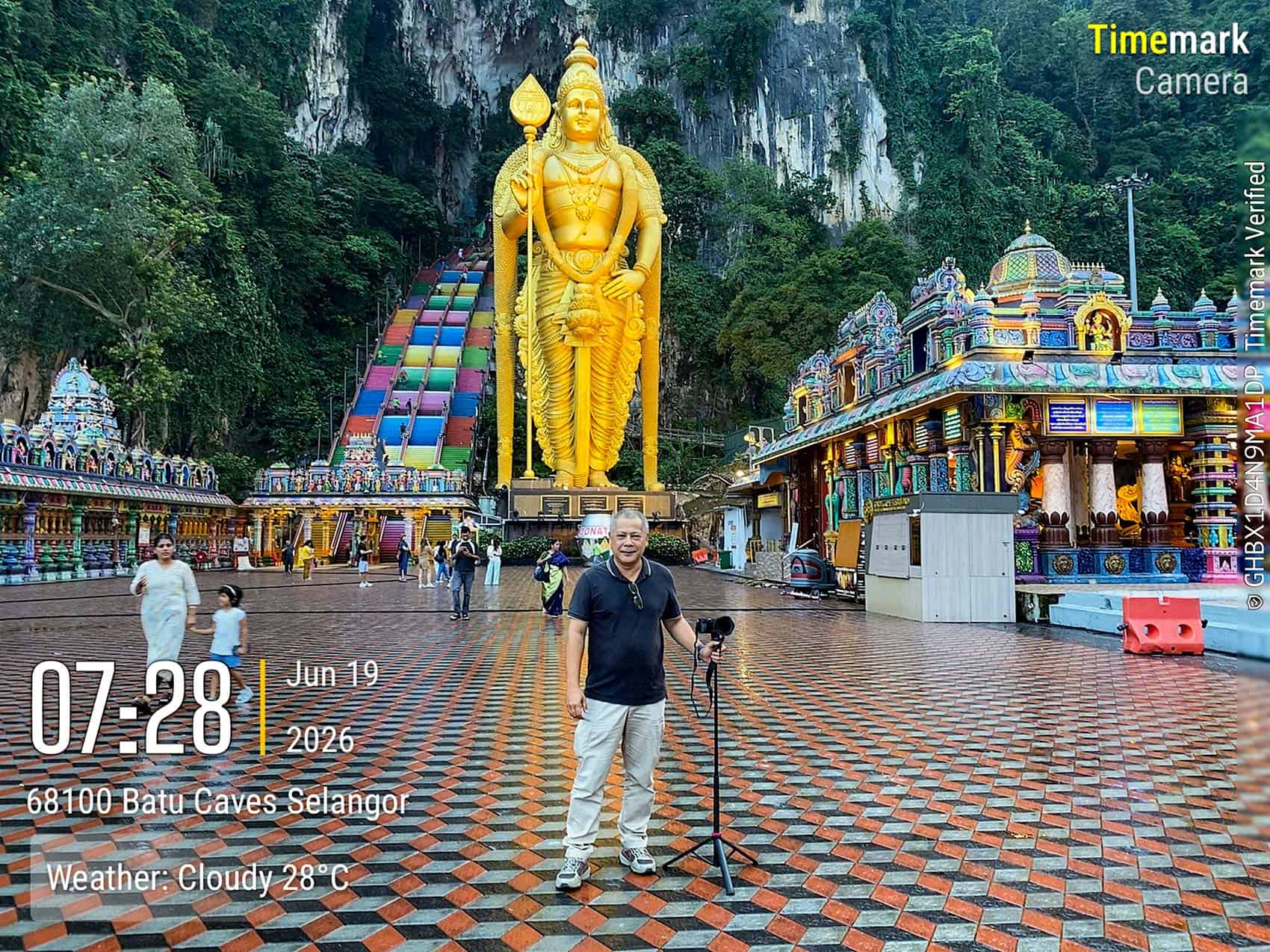 Batu Caves forecourt at 7:28 AM on 19 June 2026 — founder of We Go With Anuar conducting a field visit to verify opening hours, with Lord Murugan statue and rainbow staircase in the background