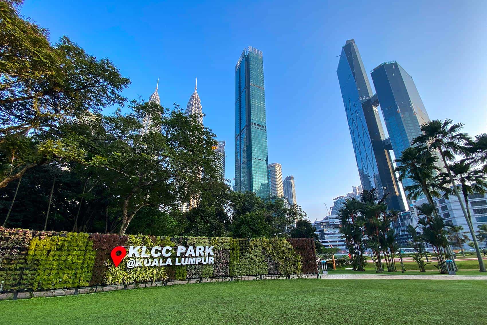 KLCC Park green lawn and living wall sign with the Petronas Twin Towers spires and Kuala Lumpur skyline visible through the trees on a clear morning