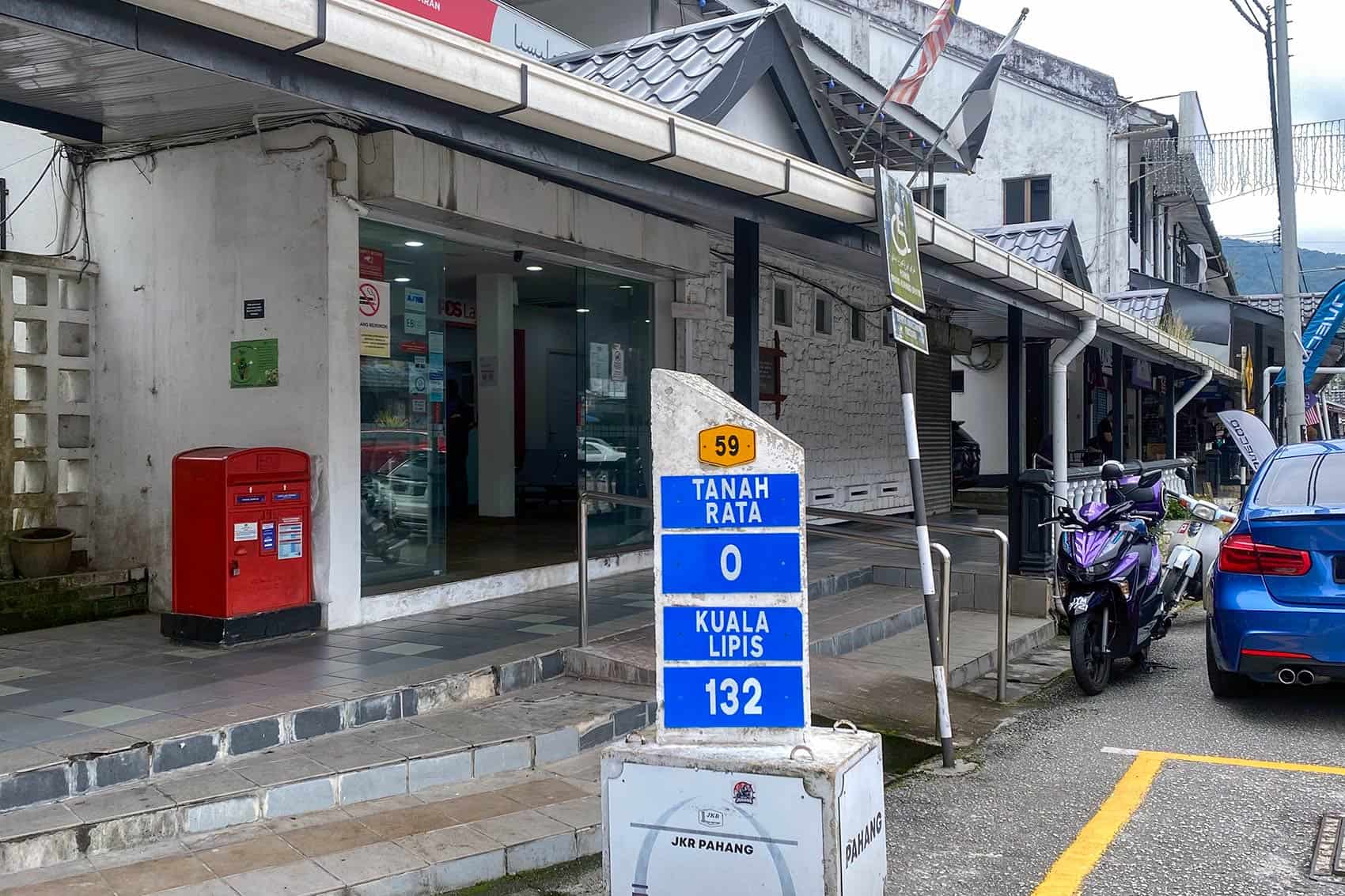 Kilometre marker at Tanah Rata town centre on Route 59 showing zero distance, with shophouses and Pos Laju office in the background — best area to stay in Cameron Highlands