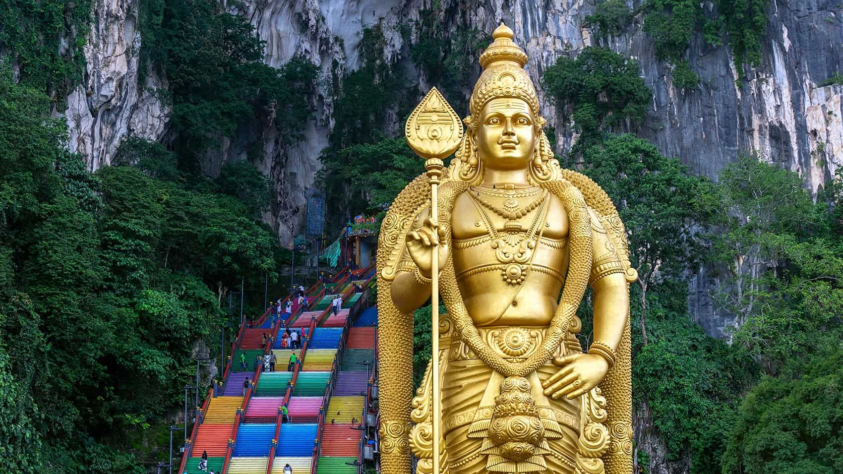 Golden statue of Lord Murugan holding the Vel spear, with Batu Caves' rainbow staircase and limestone cliffs behind it — the Batu Caves Lord Murugan statue