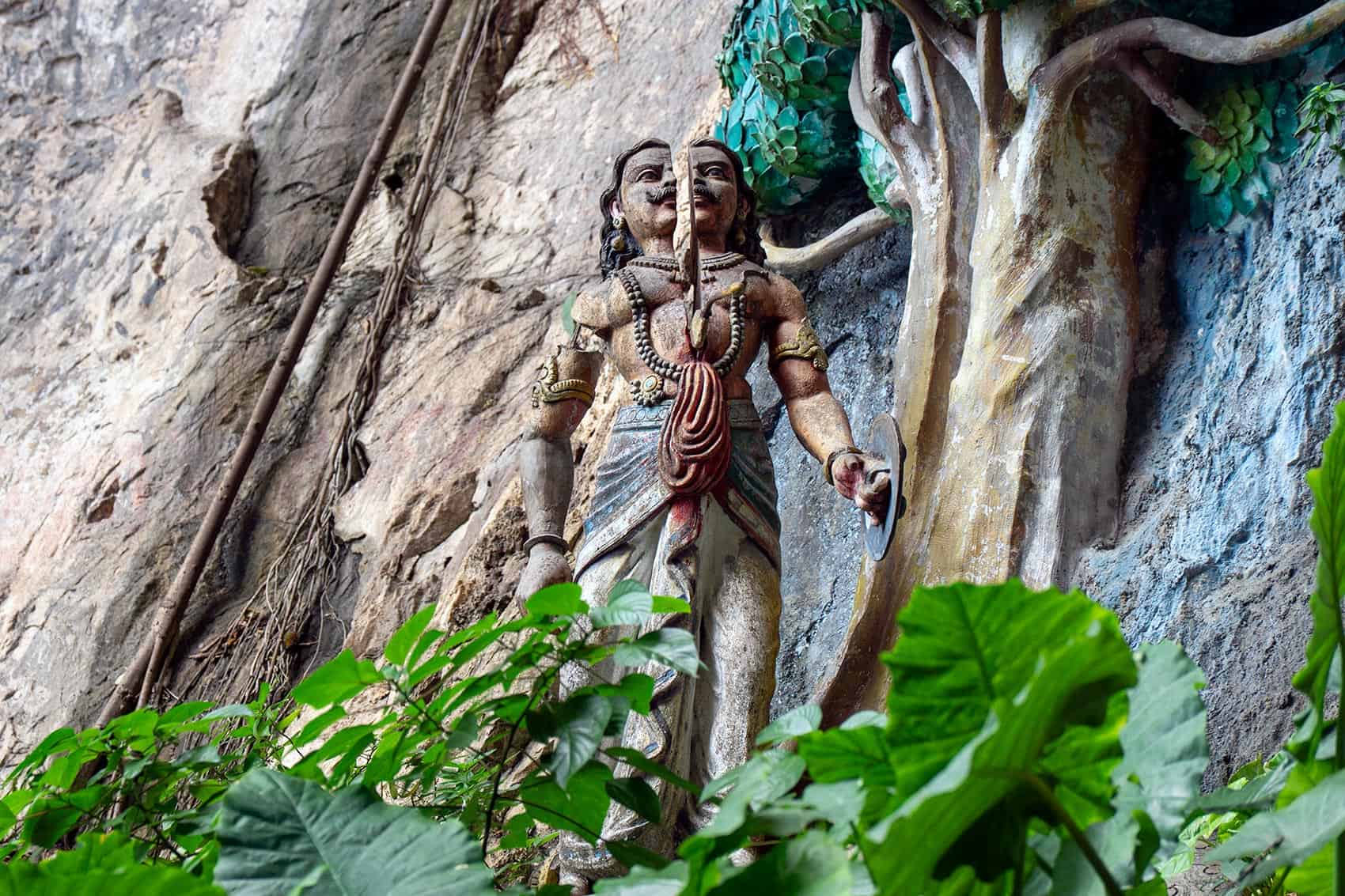 Two-faced Surapadman demon carved into the limestone cliff face at Batu Caves, depicting the moment Murugan's Vel splits him in two