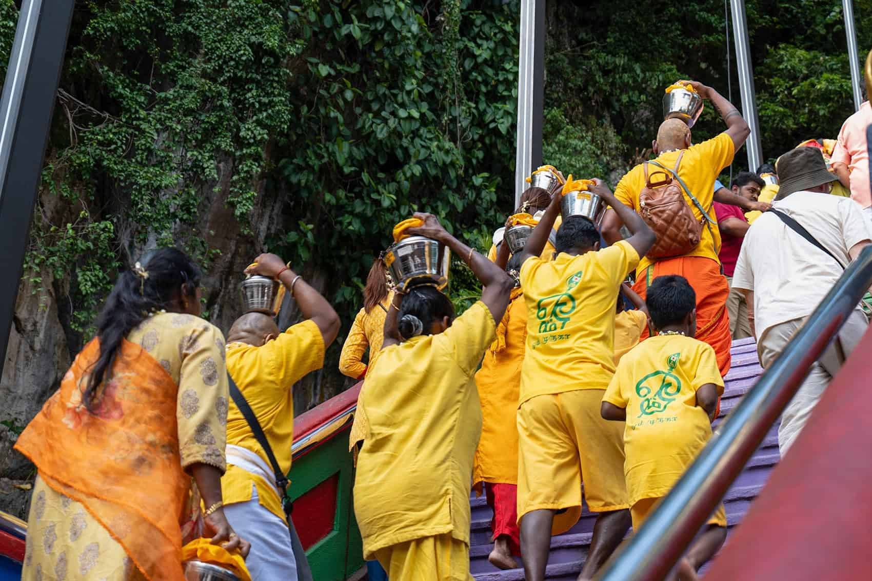 Hindu pilgrims in yellow carrying stainless steel paal kudam milk pots up the rainbow-painted steps during Thaipusam at Batu Caves